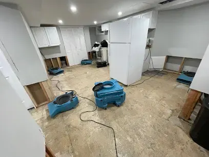 Air movers drying a basement kitchen with the lower walls cut open after water damage in Queens