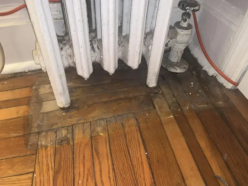 Cast-iron steam radiator and valve with water-stained oak floorboards in an Elmhurst apartment